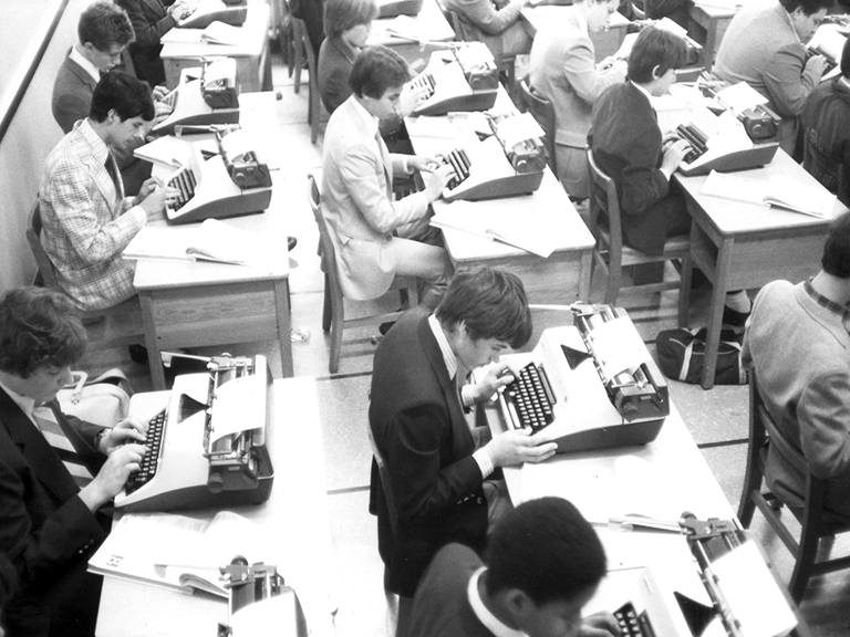 An overhead image of several young men, dressed in suits and seated at rows of desks, all learning how to type on typewriters.